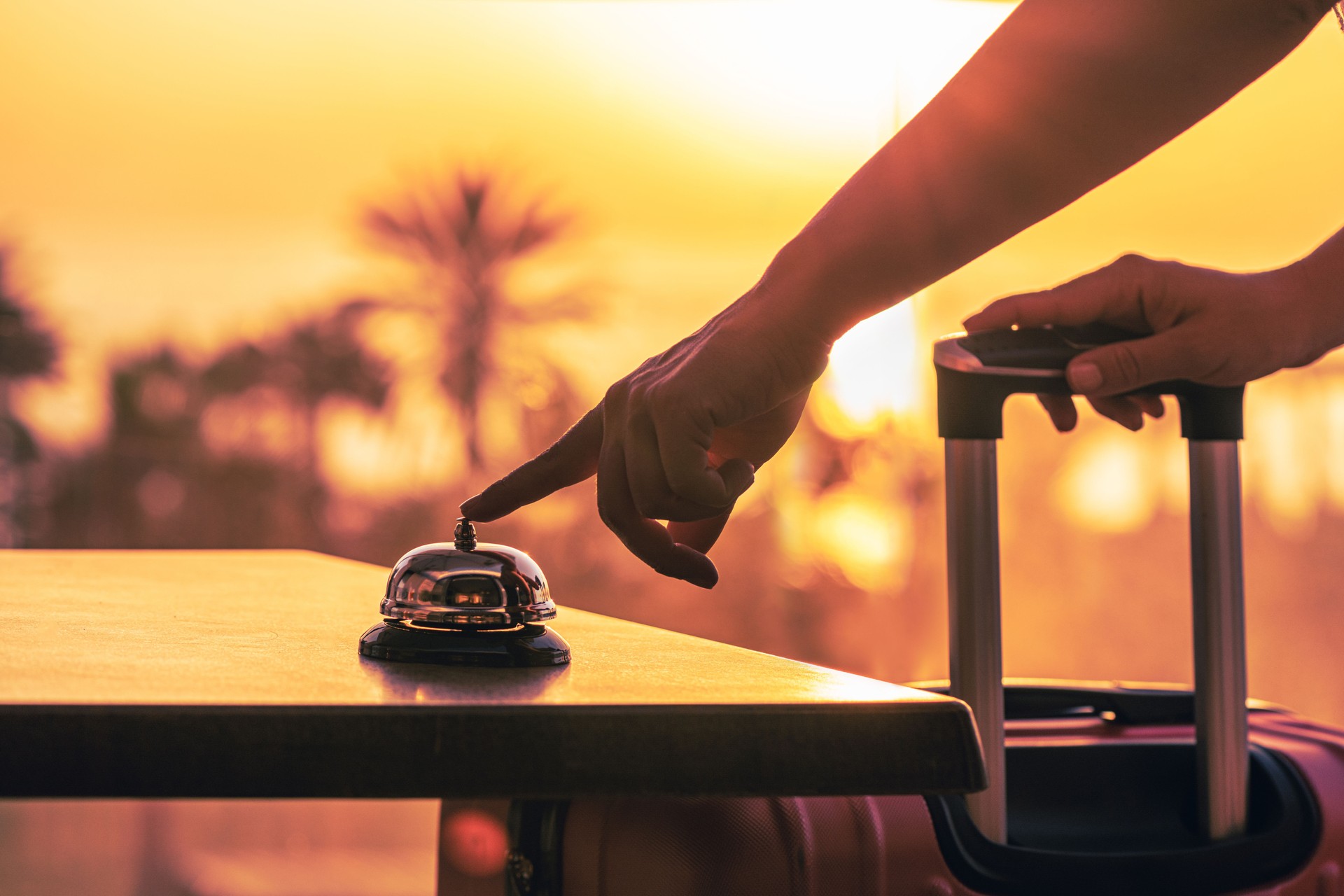 Woman with suitcase ringing hotel service bell with sea and palm tree view on sunset. Travel concept. 24-hour beach hotel front desk. Late check-out.